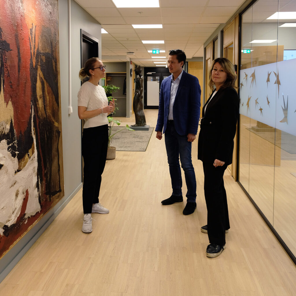 3 mennesker står utenfor et møterom, to snakker sammen, en ser i kamera